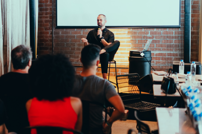 Un intervenant parle à un public dans un café, avec des murs en briques et un rétroprojecteur.